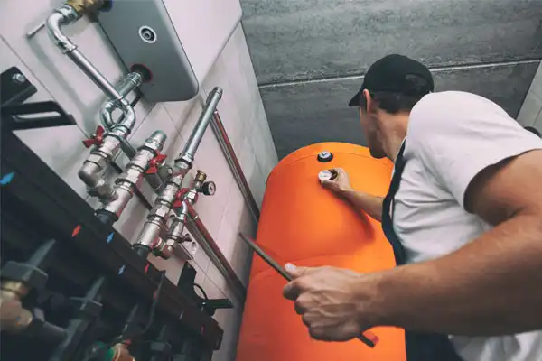 A Coast to Coast technician in a cap adjusts a gauge on an orange water heater tank in a utility room lined with pipes and valves.