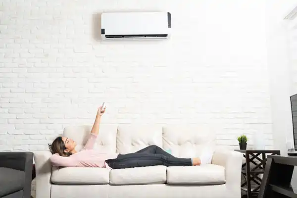 A Coast to Coast customer relaxes on her sofa in a sunlit living room, using a remote to operate her wall-mounted air conditioner.