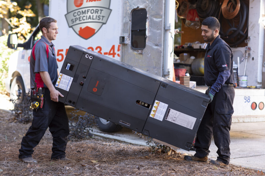 Two Coast to Coast technicians in uniform carry an HVAC unit outdoors by their service van, ready to assist a valued customer.