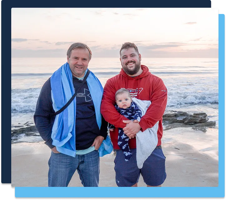 Two Coast to Coast team members stand on the beach at sunset, one gently holding a bundled infant. Waves and rocks complete the scene.